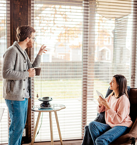 A man holds a cup of coffee and gestures while a woman seated on a chair looks at her tablet, with sunlight filtering through blinds.