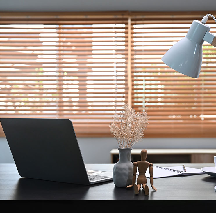 A workspace featuring a laptop, a vase with dried flowers, a wooden mannequin, and a desk lamp, with light filtering through wooden blinds.